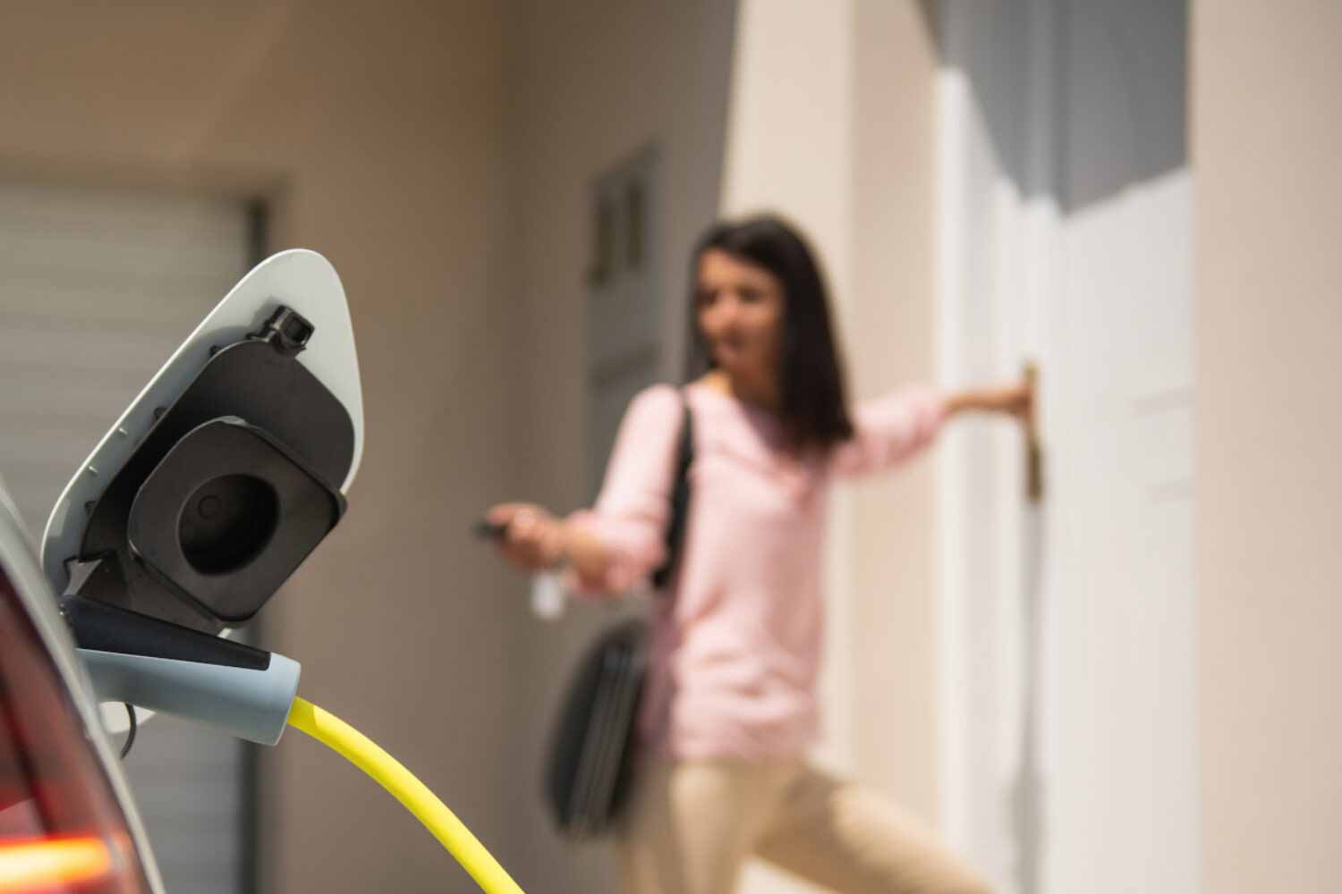 Detailansicht eines Ladekabels angeschlossen an ein Elektrofahrzeug. Verschwommen im Hintergrund eine Frau, die mit dem Funkschlüssel das Fahrzeug schließt.